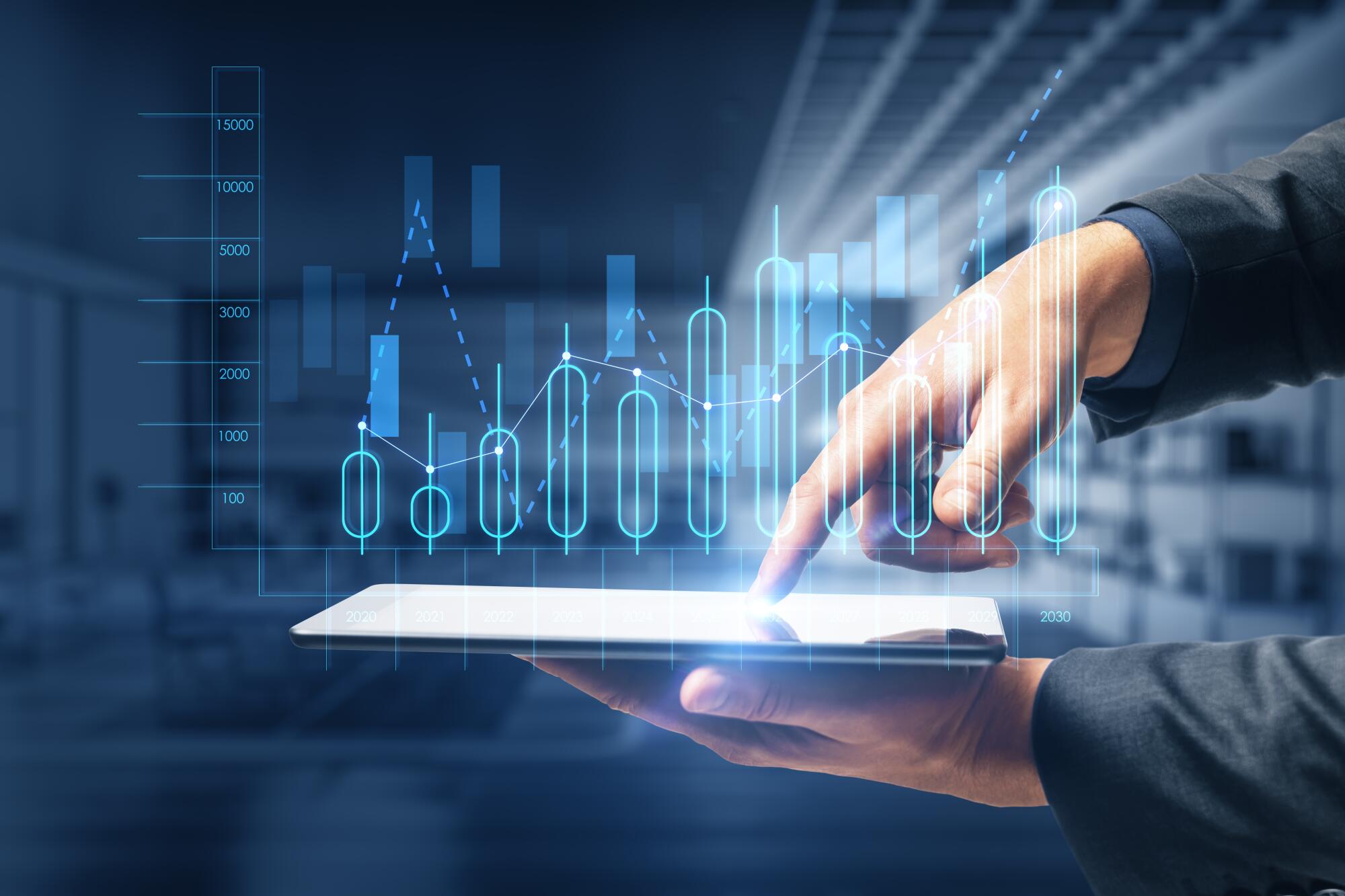 A person in a dark suit holds a white tablet while pointing at blue digital charts and graphs floating above it.